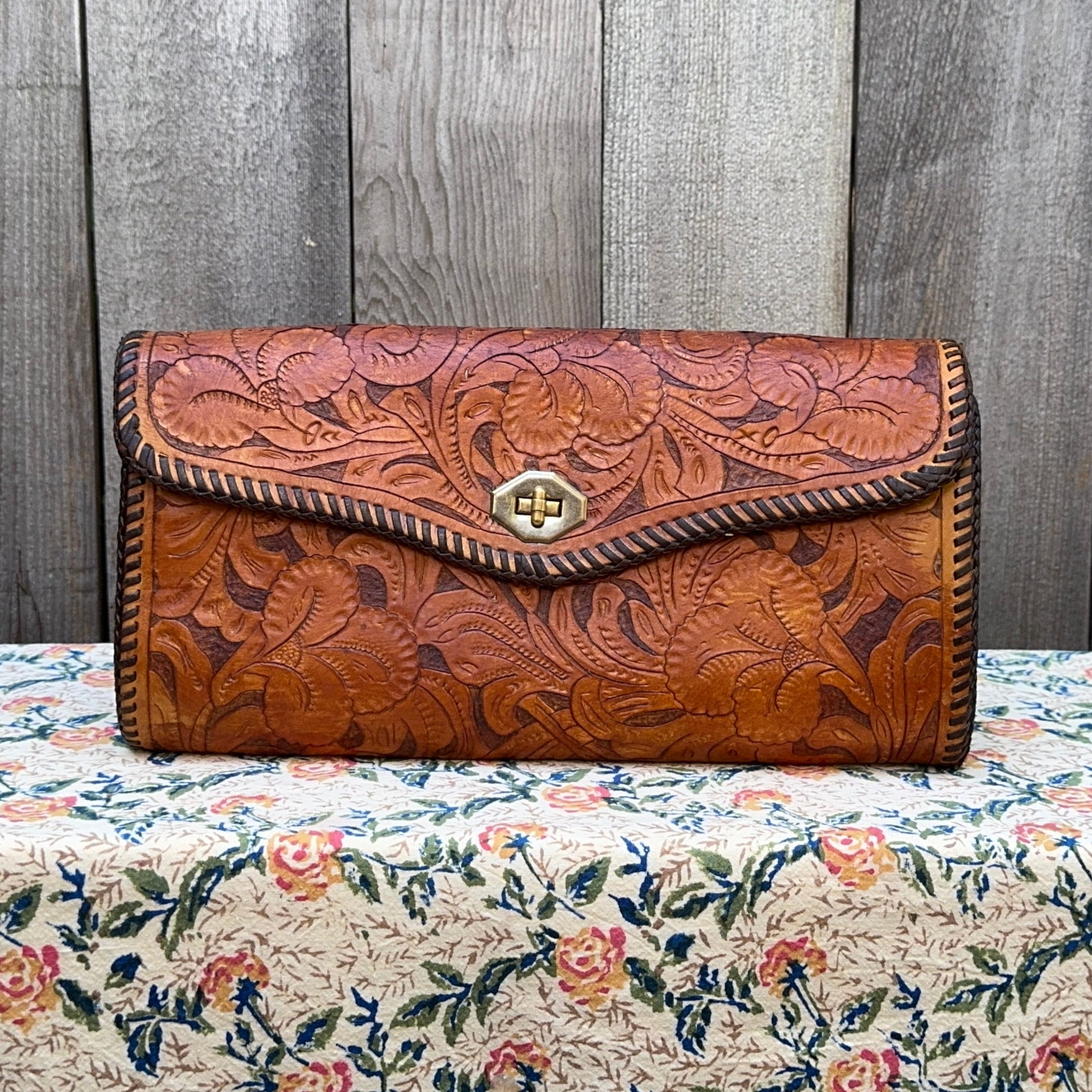 Brown leather clutch with floral patterns on a floral tablecloth against a wooden panel background