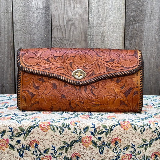 Brown leather clutch with floral patterns on a floral tablecloth against a wooden panel background